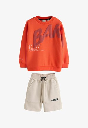 Oranžinis ilgomis rankovėmis džemperis su dideliu užrašu „BAKER“ ir smėlio spalvos šortai su raišteliais, priekiniu kišeniu ir juodu užrašu ant kairės kojos.