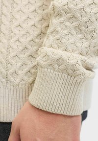 Ściśle beżowy sweter z dzianiny, z rękawem o teksturowanym wzorze diamentowym, ściągaczem oraz miękkim wełnianym materiałem. Zbliżenie na rękę i tkaninę.