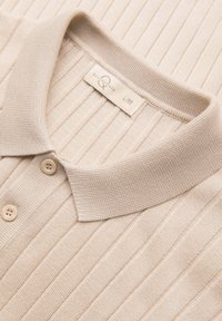 Polo en maille côtelée beige avec un col souple plié et deux boutons beiges sur la patte de boutonnage.
