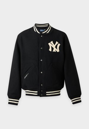 Homme portant une veste universitaire noire avec des rayures blanches et un logo, un pantalon beige à jambes larges et des chaussures noires, debout les mains dans les poches.