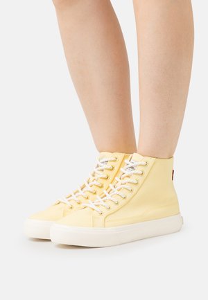 Pehmeästä keltaisesta kankaasta valmistetut korkeavartiset tennarit, joissa on valkoiset nauhat, pyöreä kärki ja kontrastinen, teksturoitu kumipohja; reunassa pieni logo.