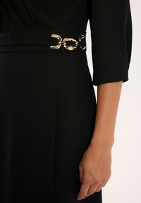 Czarna sukienka z gładkiego materiału, z rękawami o długości trzech czwartych i dekoracyjnym metalowym paskiem akcentującym talię.