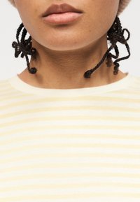 Κοντινό πλάνο του κατώτερου μέρους του προσώπου και του λαιμού ενός ατόμου με πλεγμένα μαλλιά, που φοράει πουκάμισο με ανοιχτό κίτρινο και λευκό ριγέ σχέδιο.