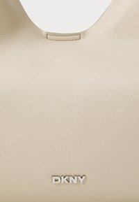Beige lærveske med en teksturert overflate, med en sirkulær åpning og en sølv "DKNY" logo nederst.