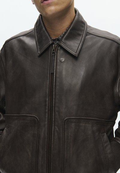 Férfi sötétbarna bőrkabátban, nagy első zsebekkel, cipzárral és patentos gallérral, mintás ing fölött.