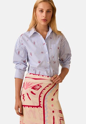 Donna con capelli biondi lunghi che indossa una camicia a righe blu con stampe rosse a tema mare e tiene una sciarpa beige con motivi rossi e rosa.