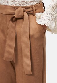 Pantalons taille haute en daim marron avec un détail de ceinture nouée, présentant une texture lisse et des poches latérales, complétés par un chemisier en dentelle.