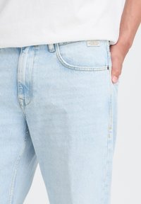 La personne porte un jean en denim bleu clair et une chemise blanche, la main droite reposant dans la poche avant droite.