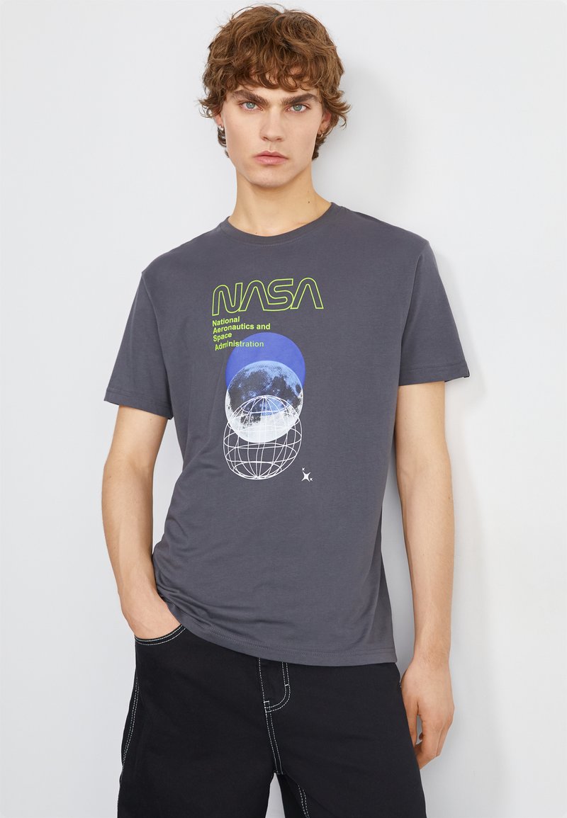 T-shirt gris avec un graphique NASA ; présente des images de planètes bleues et blanches et du texte vert. Fait en coton avec des manches courtes et un col rond.