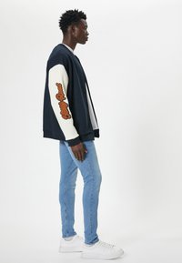 Laivasilta sininen, ylisuuri takki valkoisilla hihoilla, jossa on oranssia brodeerattua tekstiä. Vaaleansiniset slim-fit farkut ja valkoiset lenkkarit täydentävät asun.