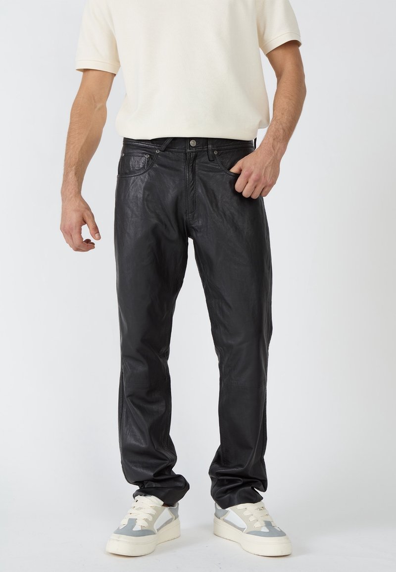 Homme portant un pantalon en cuir noir, des baskets blanches et grises, et une chemise blanche à manches courtes sur un fond clair uni.