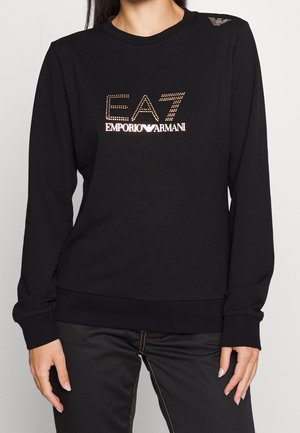 Chemise noire à manches longues avec logo EA7 Emporio Armani clouté sur le devant, portée avec un pantalon noir aux coutures visibles.