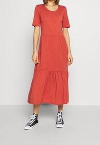 Rotes, kurzärmliges Midikleid mit tailliertem Oberteil und gestuftem Rock. Trägt schwarze High-Top-Sneaker. Weicher, glatter Stoff.