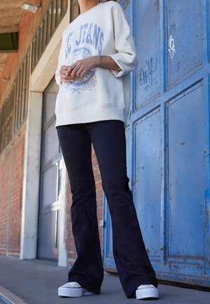 Sudadera crema con texto gráfico azul, combinada con pantalones acampanados negros y zapatillas blancas de plataforma. El fondo presenta una puerta azul.