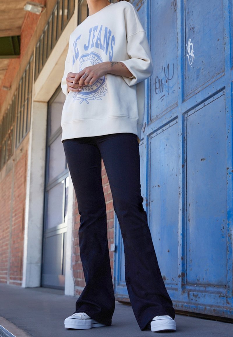 Sudadera crema con texto gráfico azul, combinada con pantalones acampanados negros y zapatillas blancas de plataforma. El fondo presenta una puerta azul.
