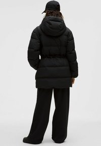 Giacca a vento nera con cappuccio, vita aderente e texture trapuntata. Caratterizzata da maniche lunghe e un taglio leggermente allungato, abbinata a pantaloni neri.