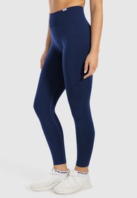 Granatowe legginsy high-waisted wykonane z elastycznego materiału. Charakteryzują się gładką teksturą i kontrastowymi szwami. Połączone z białymi butami sportowymi.