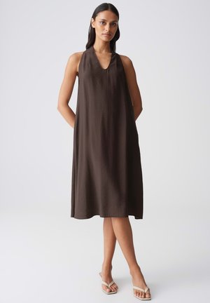 Donna in piedi con le mani dietro la schiena, indossa un vestito senza maniche marrone scuro, lungo fino al ginocchio, e sandali aperti chiari su sfondo bianco.