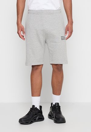 Mann trägt hellgraue kniehohe Shorts mit schwarzem Text am linken Bein, weiße Sportsocken und schwarze Sportschuhe.