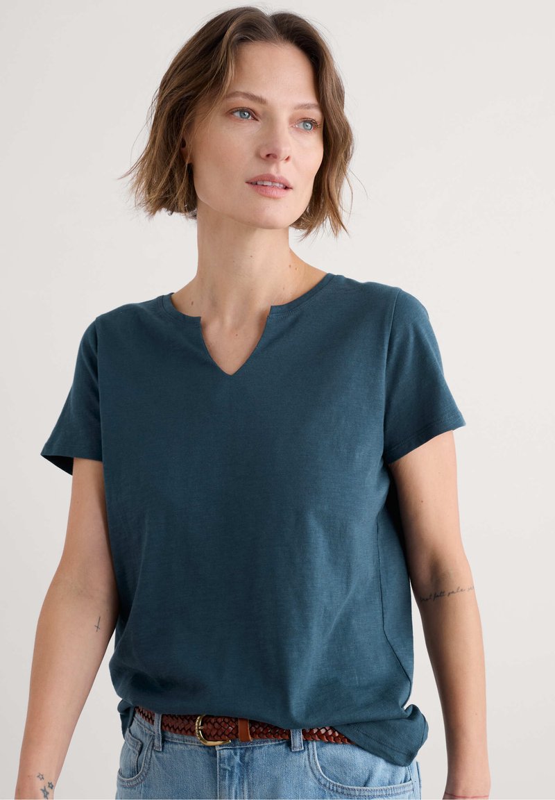 Tyrkysové tričko s krátkými rukávy, výstřihem do V a volným střihem. Vyrobené z měkké látky s jemnou texturou a bočními rozparky pro lepší pohyblivost.