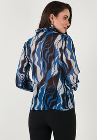 Sheer blouse featuring a wavy pattern in shades of blue, brown, and cream. It has long sleeves and a ruffled collar detail.