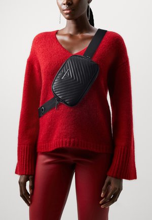 Kobieta w czerwonym swetrze i spodniach z czarną pikowaną torbą na ramię z szerokim paskiem przewieszonym skośnie przez klatkę piersiową.