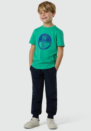 Ragazzo con capelli biondi che indossa una maglietta verde "North Sails", pantaloni blu navy, sneakers bianche, in piedi con le mani in tasca, sorridente.