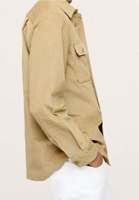 Mand iført en beige kordfløjlsskjorte med knaplukket brystlomme, hånd i lommen, stående mod en ensfarvet lys baggrund.