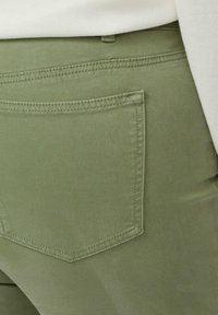 Gros plan sur la poche d’un pantalon vert olive avec des coutures apparentes et une partie d’une chemise blanche rentrée.
