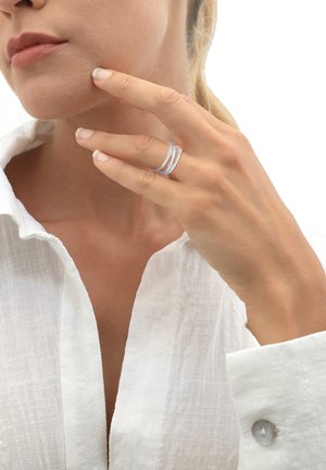Silberne Ringe, übereinander gestapelt an den Fingern, vor einem weißen Leinenhemd. Glatte, polierte Oberfläche mit einem minimalistischen Design. Natürliche Nägel sichtbar.