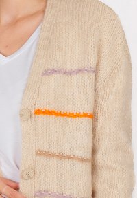 Ripr close-up di un cardigan lavorato a maglia beige con strisce arancioni, viola e marrone chiaro, indossato su una maglietta bianca a scollo a V.