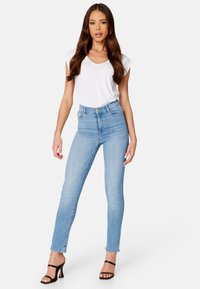 Jasnoniebieskie spodnie z wysokim stanem o slim fit, zestawione z białą koszulką z dekoltem w serek. Modelka ma na sobie czarne sandały na wysokim obcasie.