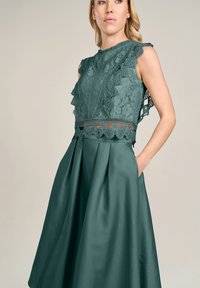 Grünes Kleid mit einem Spitzenoberteil und Rüschendetails sowie einem plissierten Satinrock. Der Stoff hat eine glatte Textur und einen subtilen Glanz.