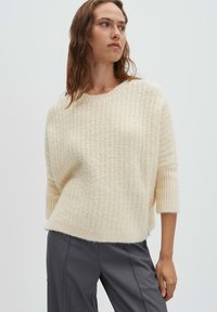 Cremefarbener, strukturierter Strickpullover mit rundem Ausschnitt und Dreiviertelärmeln, kombiniert mit grauen Hosen. Weiches, gemütliches Aussehen.