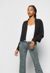 Camisa preta de botão com mangas longas sobre uma t-shirt branca, combinada com jeans flare desgastados em azul com costuras diagonais.