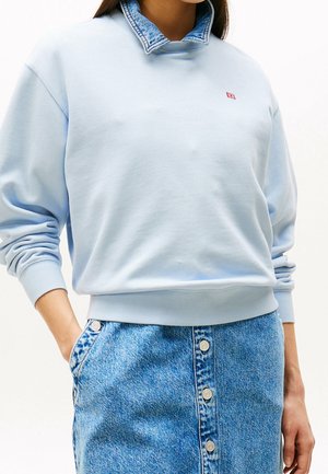 Kobieta ubrana w jasnoniebieską bluzę z kapturem na kołnierzyku z denimu i zapinaną na guziki dżinsową spódnicę, ręce w kieszeniach.