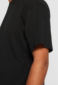 Gros plan d'une personne portant un t-shirt noir uni à manches courtes, montrant l'épaule, la manche et le haut du bras.