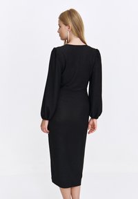 Schwarzes Kleid mit langen Puffärmeln, tailliertem Schnitt und Midi-Länge. Aus strukturiertem Stoff mit einem subtilen Glanz.