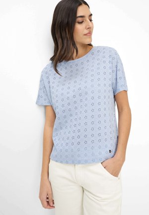 Frau mit dunklen Haaren, die ein hellblaues Kurzarmtop mit Lochmuster und cremefarbene Hosen trägt, schaut nach unten und hat eine Hand in der Tasche.