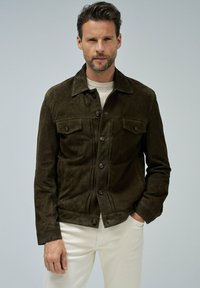 Homme portant une veste en daim marron foncé sur un pull blanc et un pantalon blanc, se tenant avec une main dans la poche contre un fond uni.