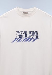 Weißes Baumwoll-T-Shirt mit dem Wort "NADA" in einer blauen, strukturierten Schrift mit Schatteneffekt, Rundhalsausschnitt und kurzen Ärmeln.