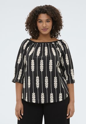 Kobieta z kręconymi włosami, ubrana w czarną bluzkę odkrywającą ramiona z pionowymi kremowymi wzorami liści i czarne spodnie, stojąca na jednolitym tle.