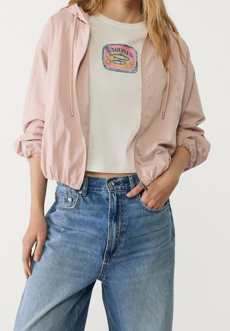 Jasnoróżowa kurtka typu bomber z elastycznymi ściągaczami na rękawach, noszona na białej, krótkiej koszulce z kolorowym nadrukiem sardynek, zestawiona z jeansami z wysokim stanem.