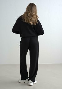 Svart oversized hettegenser kombinert med løs svarte bukser, begge i et mykt stoff. Hvite sneakers med minimale detaljer fullfører antrekket.