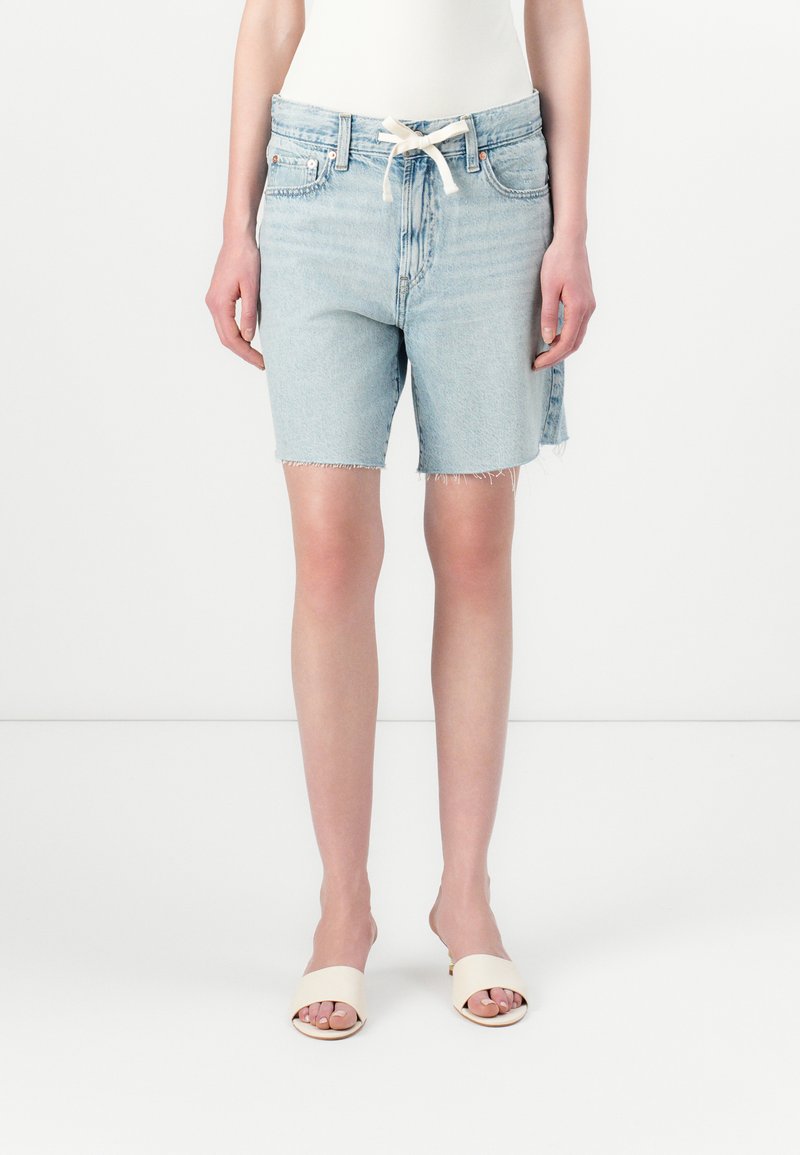 Ljusblå denimshorts med fransiga kanter, hög midja och dragsko. Bärs med neutrala öppna sandaler.