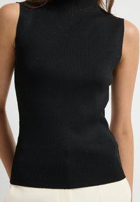 Top senza maniche in maglia a coste nera con sottili punti luccicanti indossato da una persona, abbinato a pantaloni di colore chiaro.