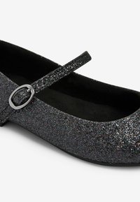 Bailarinas negras con purpurina, punta redondeada, textura similar a la satén y suela plana. Presentan una correa con detalle de hebilla plateada.