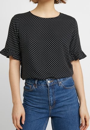 Persoon draagt een zwarte korte mouw top met witte stippen en geruite mouwen, gecombineerd met high-waisted blauwe jeans.