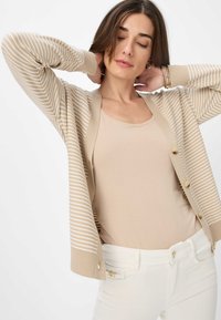 Beige gestreifter Cardigan mit langen Ärmeln, verziert mit Knöpfen und einer weichen Textur, über einem schlichten beigen Oberteil getragen. Kombiniert mit weißen Hosen.