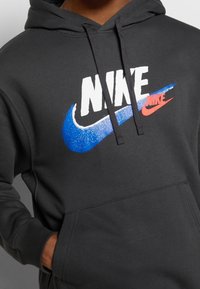 Tmavě šedá mikina s kapucí s velkým bílým logem "NIKE" v modrých a červených akcentů, vyrobená z měkké, strukturované látky s přední kapsou.